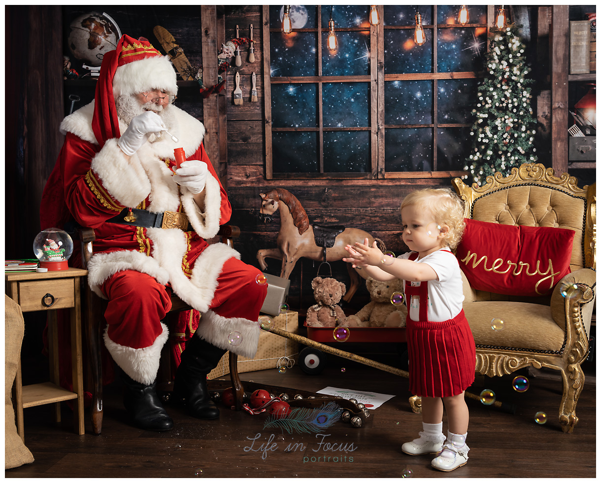 Blowing Bubbles with Santa Life in Focus Portraits Christmas photoshoots Photo of Santa blowing bubbles to little girl in red dress Christmas photoshoot at Life in Focus Portraits child photographer Helensburgh