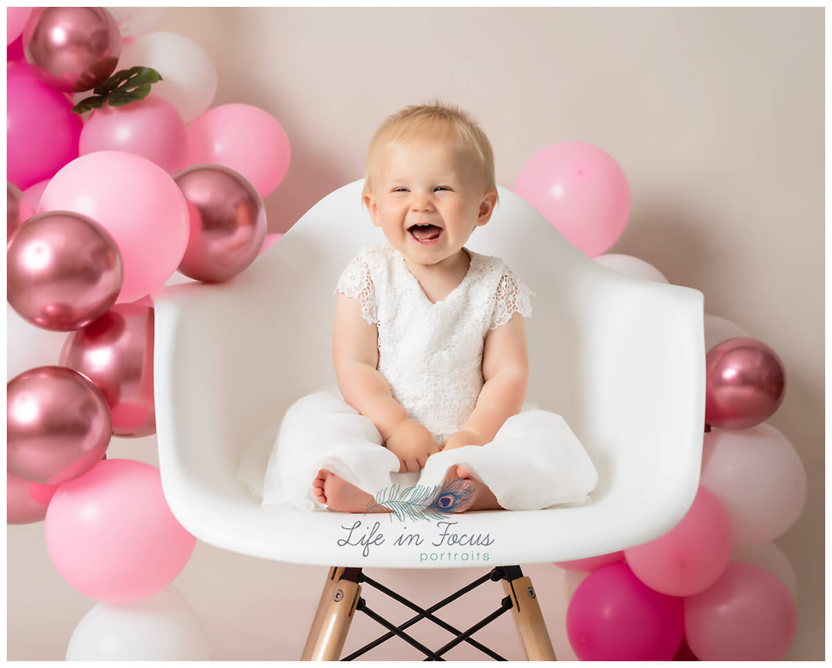 photo of laughing baby girl sitting on chair surrounded by pink balloons 1st Birthday photoshoot Rhu