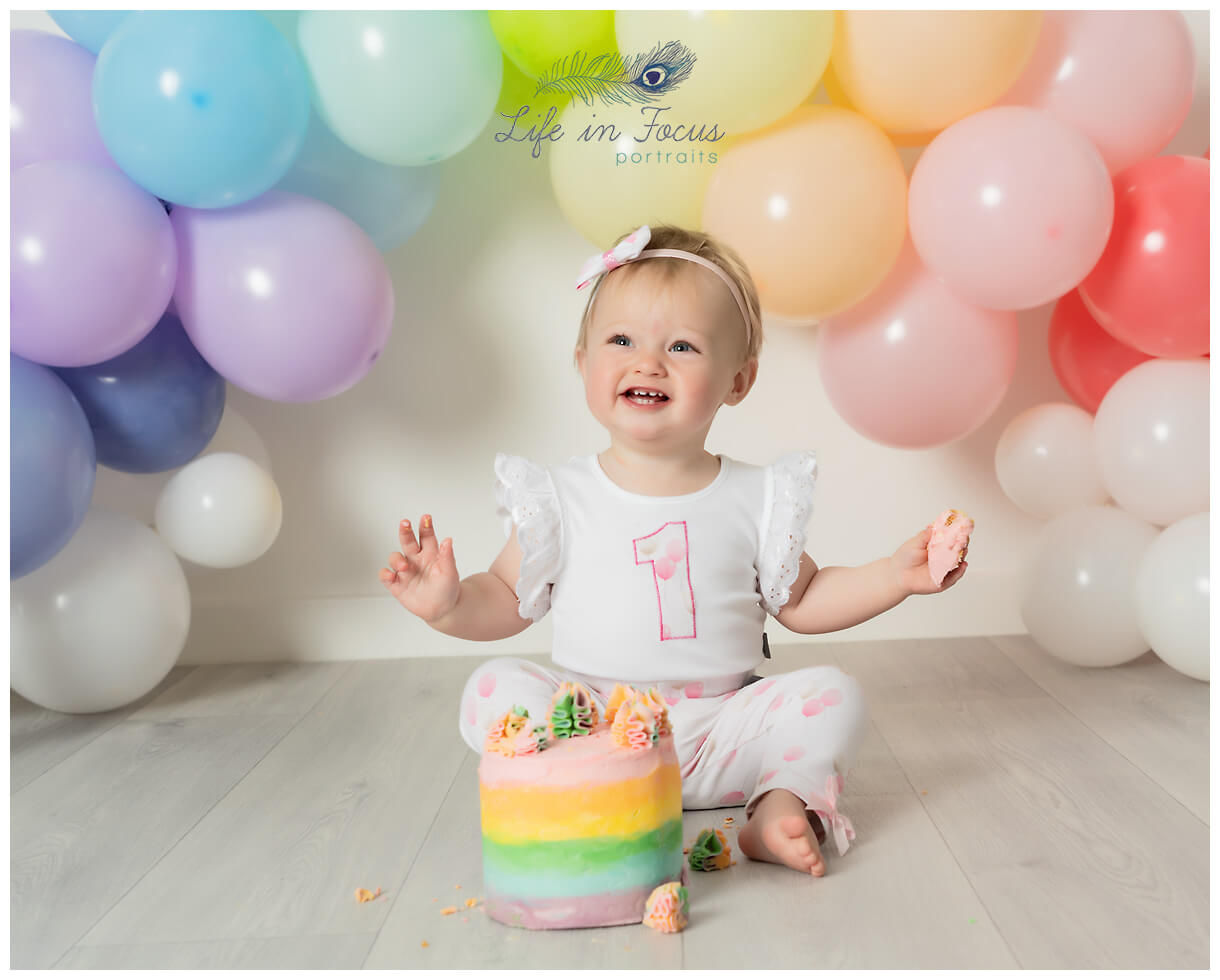 photo of smiling baby girl with rainbow cake 1st Birthday studio photographer