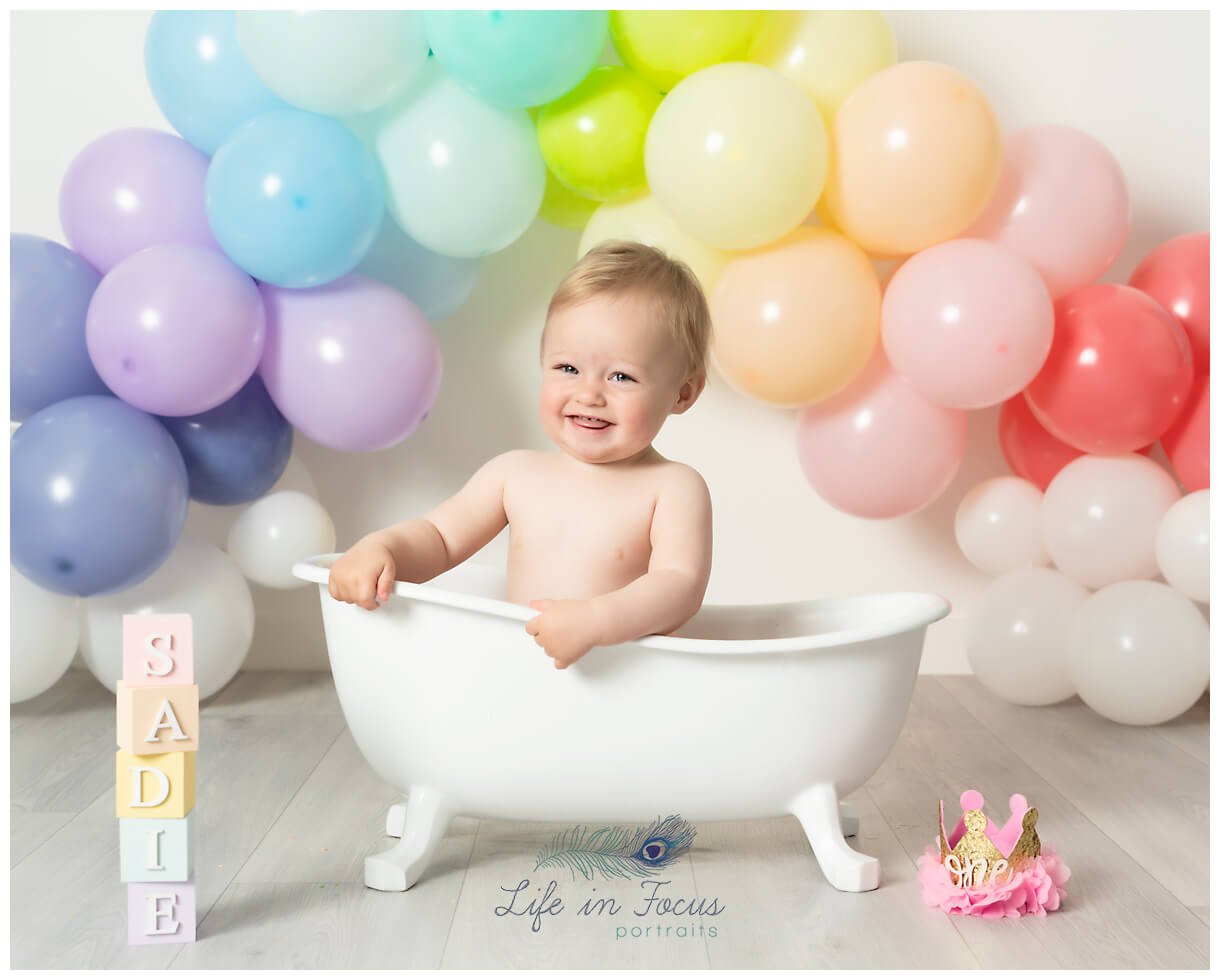 photo of smiling baby girl in bathtub with rainbow balloons smash and splash photographer Helensburgh